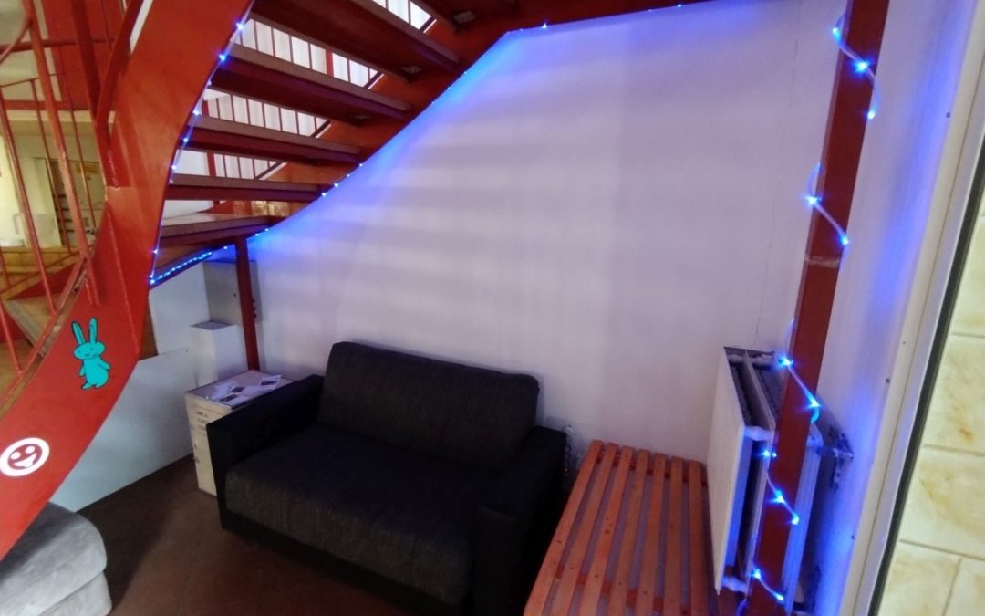 Eine Couch mit blauem Lichterschlauch steht unter einer roten Treppe.